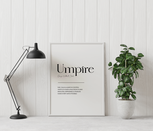 A framed typographic wall print reading 'Umpire' with smaller text below, leaning against a white paneled wall beside a black desk lamp and a potted green plant.